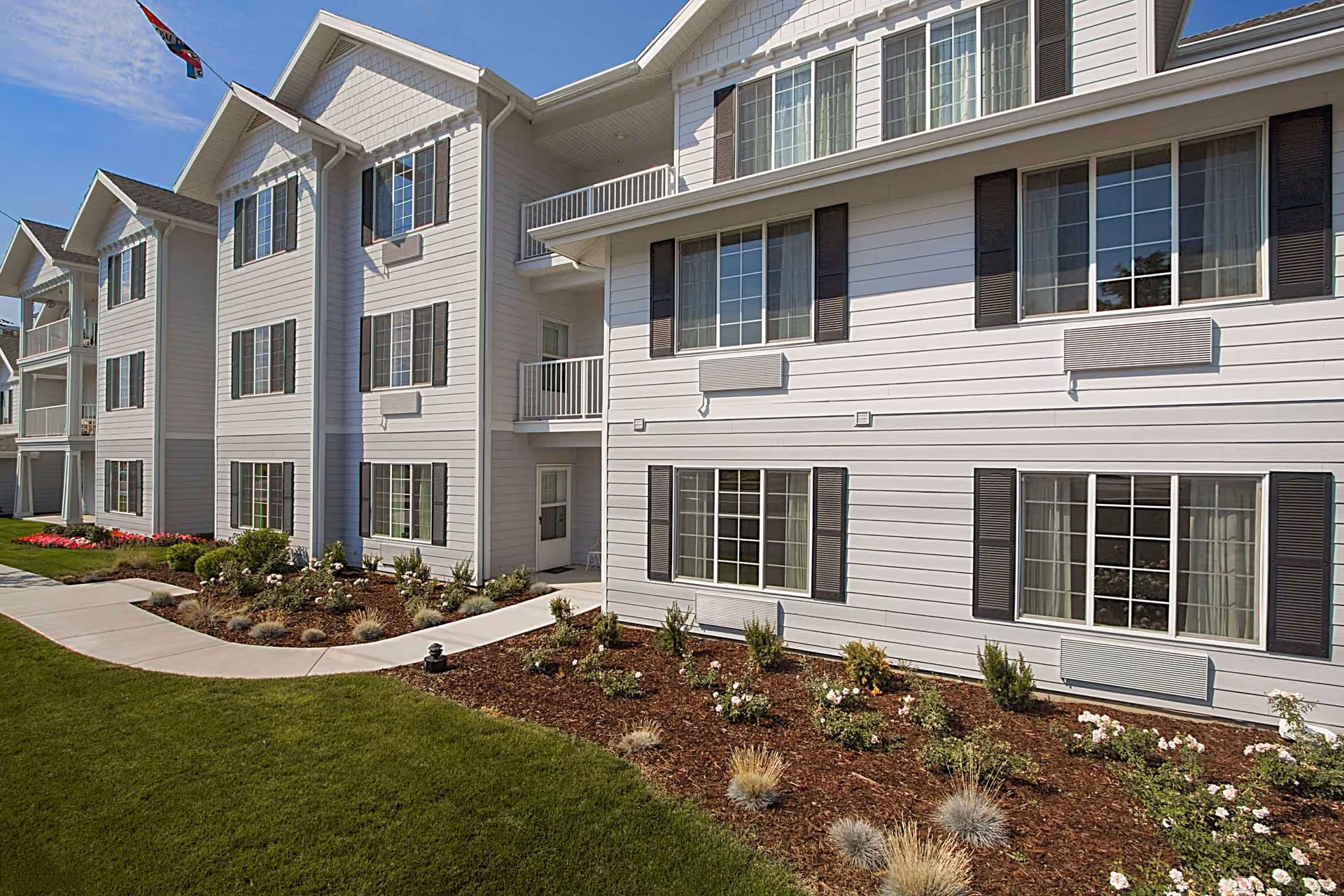 Exterior view of a multi-story residential building with white siding and dark shutters. The building has multiple large windows and small balconies. There is a well-maintained garden with various plants and flowers along a curved concrete walkway, and a green lawn in the foreground under a clear blue sky.