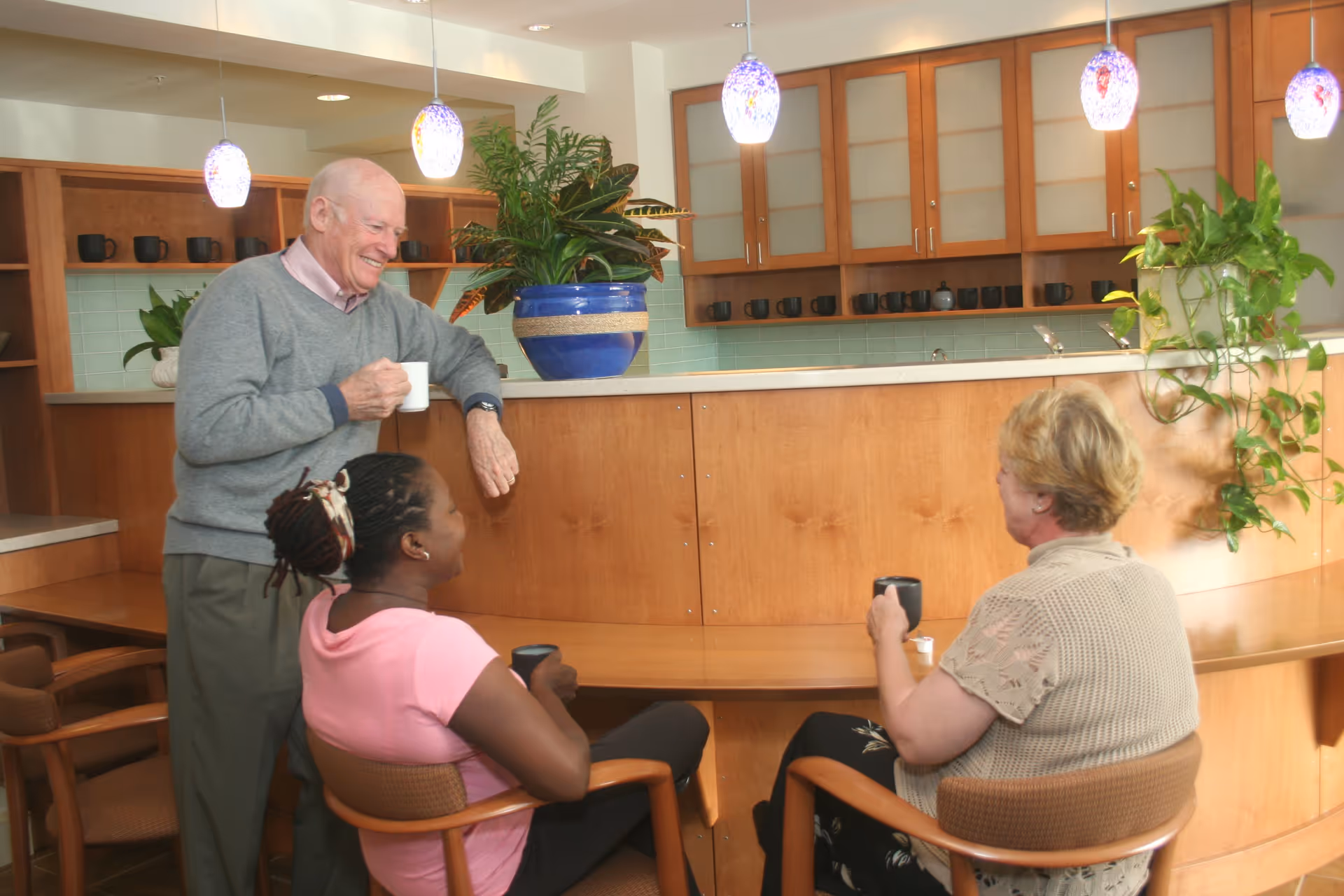 Three residents chat and drink coffee around a wooden counter in a bright communal kitchen/dining area with pendant lights and potted plants.
