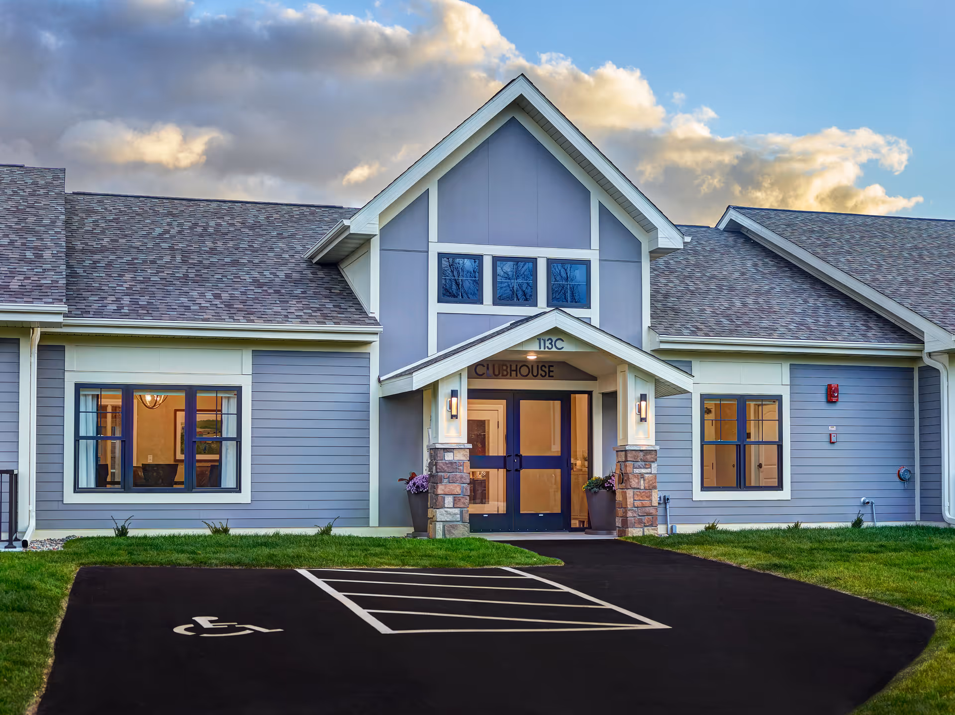 Exterior front of a clubhouse building with double entry doors, windows, entryway columns, and a nearby handicapped parking space.