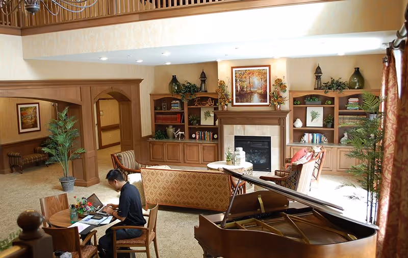 A spacious and well-lit living room area with a grand piano in the foreground, a man sitting at a small round table working on a laptop, and comfortable seating including a sofa and armchairs arranged around a fireplace with built-in wooden shelves decorated with books, plants, and artwork.