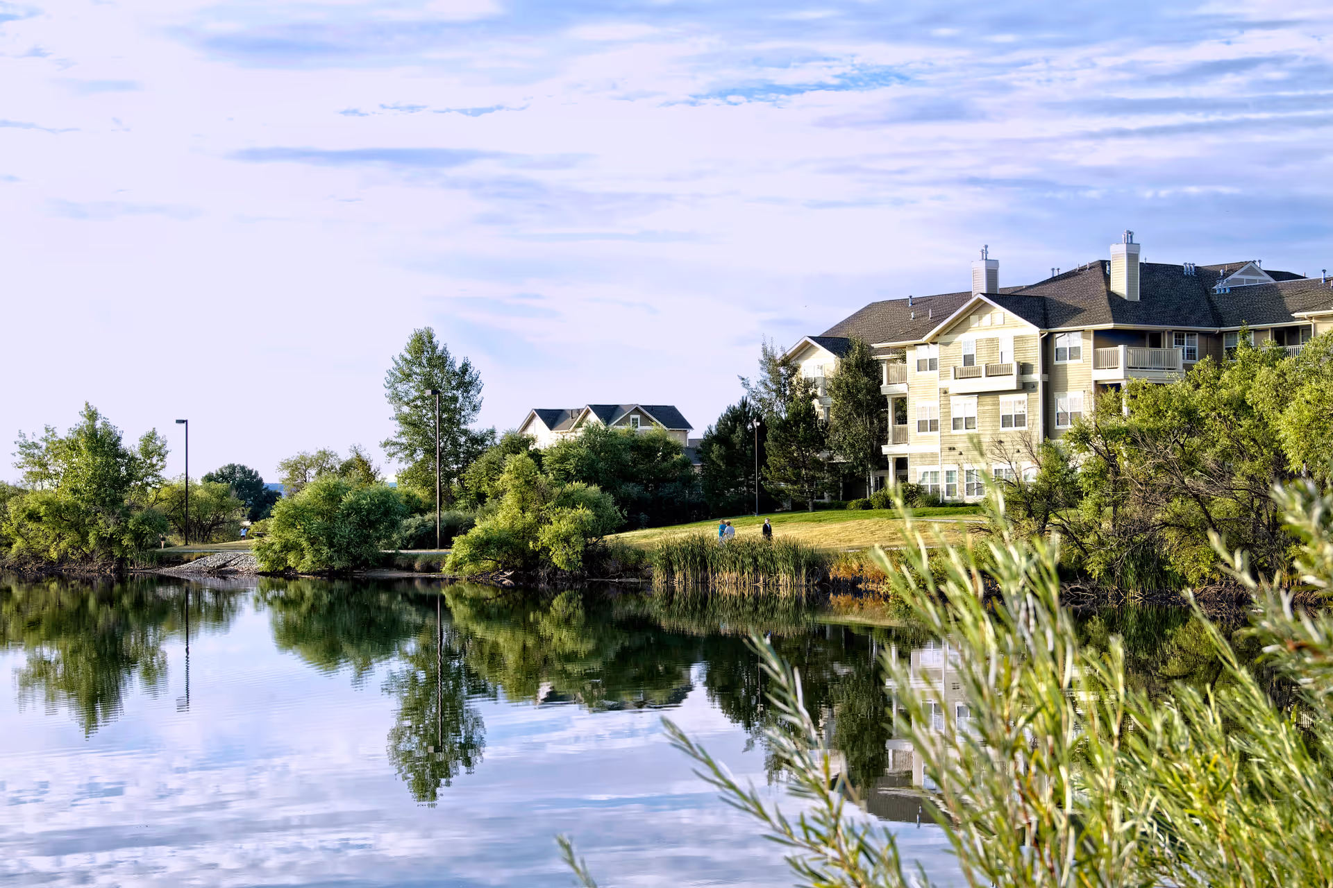 A lakeside view of a multi-story residential building reflected in calm water with trees and grassy shoreline.
