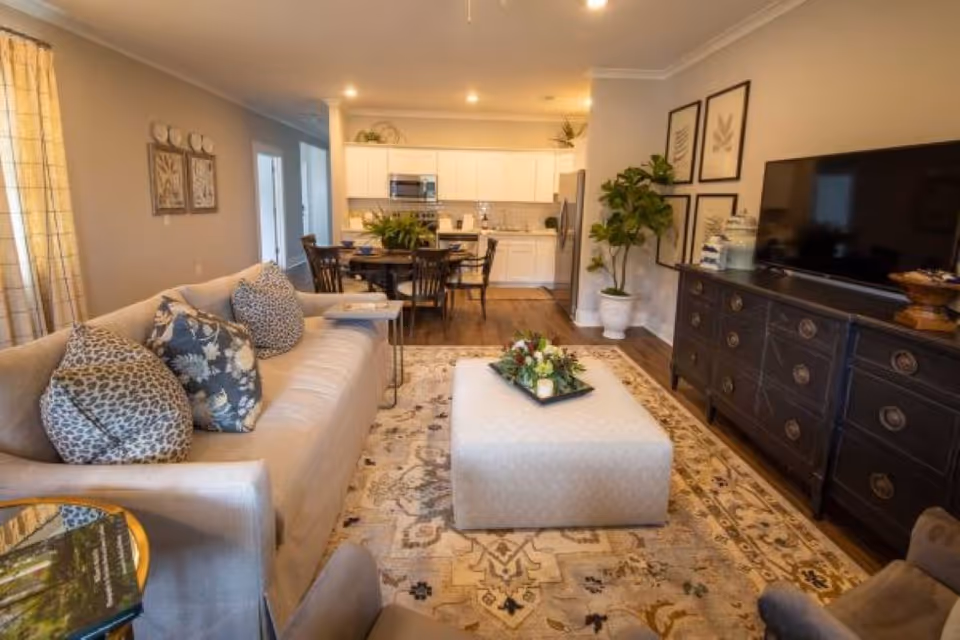 A cozy living room area with a beige sofa adorned with patterned cushions, a large ottoman with a decorative tray, a dark wooden dresser with a flat-screen TV on top, and framed botanical artwork on the wall. In the background, there is a dining table with chairs and a kitchen with white cabinets and stainless steel appliances.