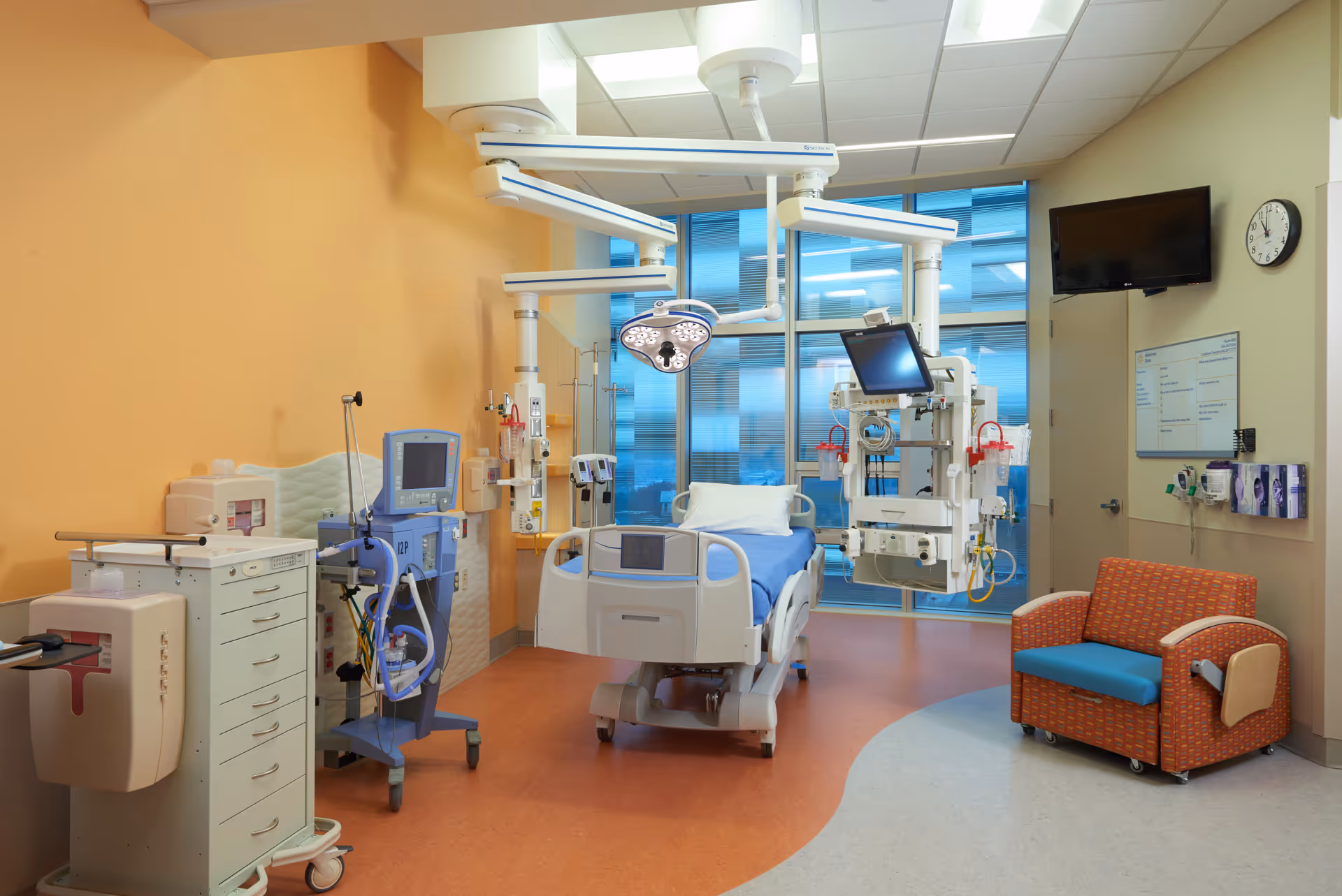 A brightly lit surgical suite with a hospital bed, anesthesia equipment, monitors, and overhead lights.
