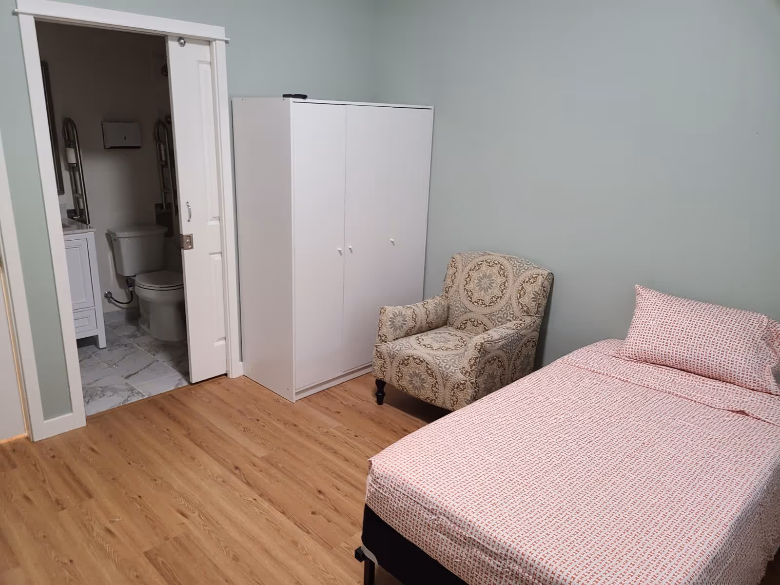 A bedroom with light green walls and wood flooring featuring a single bed with pink patterned bedding, a patterned armchair, and a white wardrobe. An open door reveals a bathroom with a toilet and a sink.