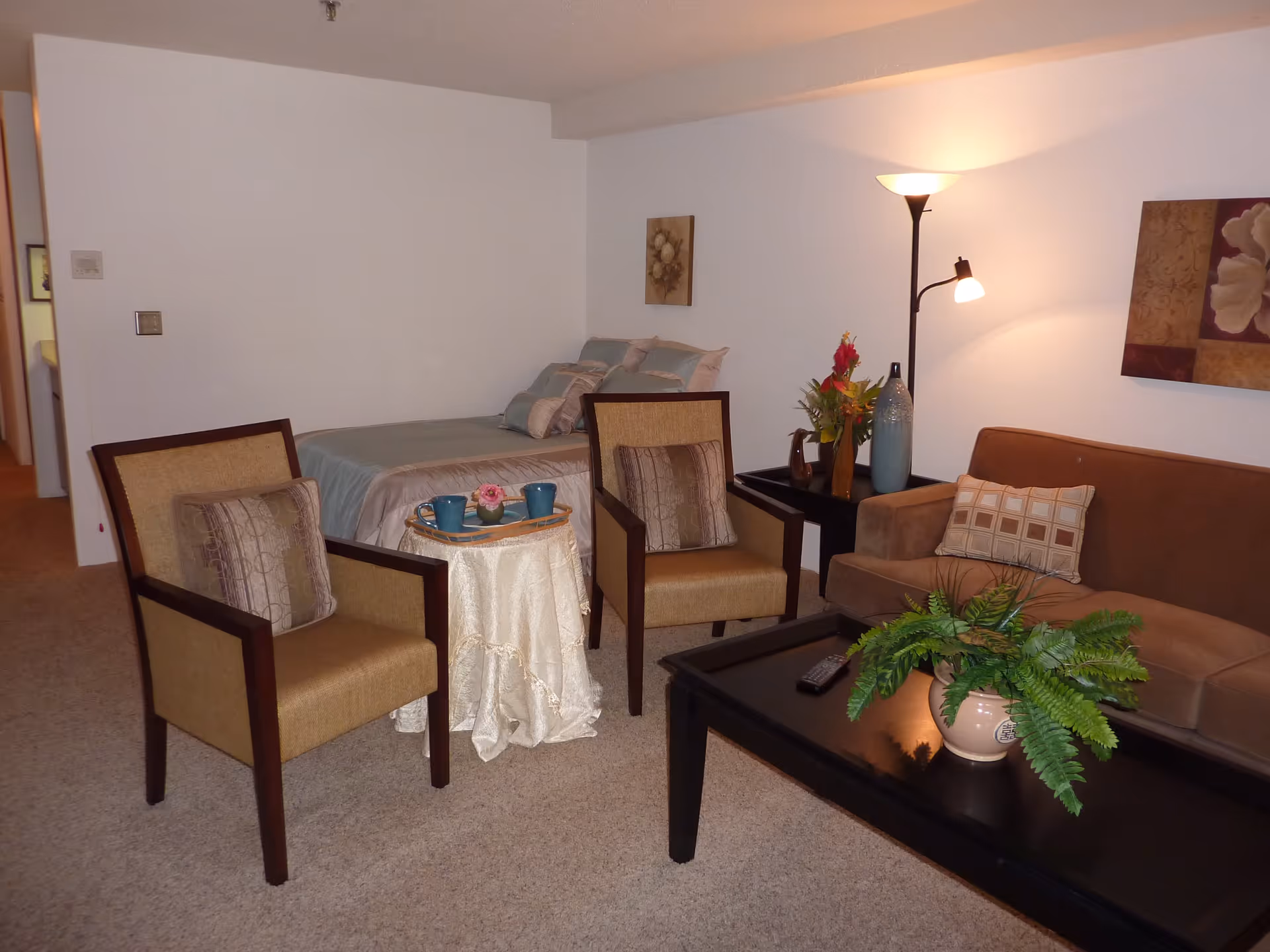 A cozy room with a bed in the background, two beige armchairs with patterned cushions, a small round table covered with a white cloth holding a tray with two blue mugs and a small flower, a brown sofa with a patterned cushion, a black coffee table with a green potted plant and a TV remote, a side table with a vase of flowers and a tall floor lamp providing warm light, and two framed floral artworks on the walls.