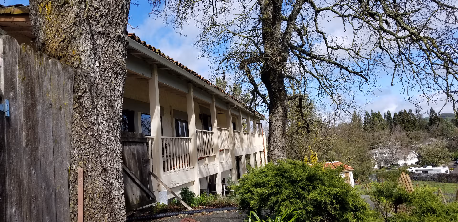 Two-story beige retirement home exterior with balconies, trees, and a wooden fence along a hillside.