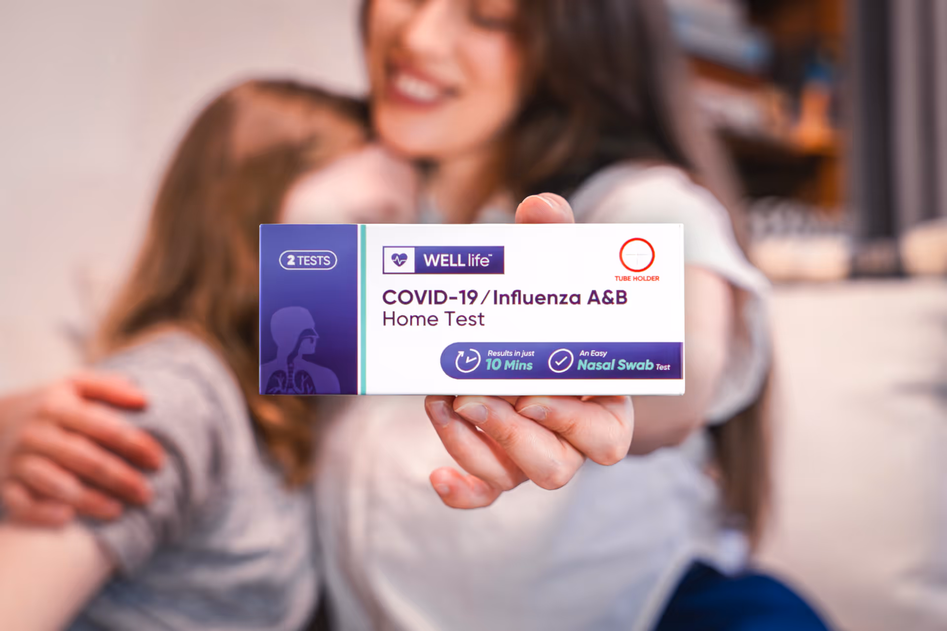 A hand extends a COVID-19/Influenza A&B home test box toward the camera, background figures embrace.