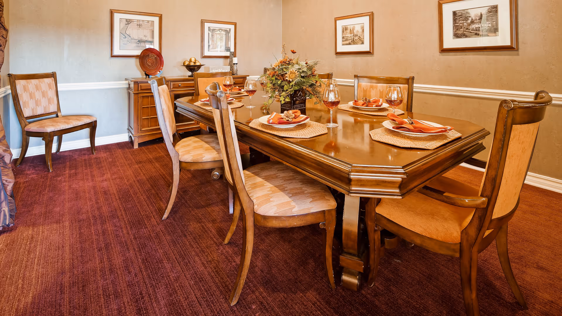 A formal dining room with a polished wooden dining table set for six people. The table has placemats, folded napkins, plates, and wine glasses. There is a floral centerpiece on the table. The room has beige walls with framed artwork and a wooden sideboard against the wall. The floor is covered with a reddish-brown carpet.