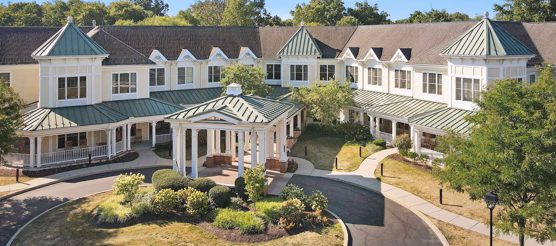 Front exterior of a two-story senior living facility with green metal roofs, a covered porte-cochere, wraparound porch and landscaped circular driveway.
