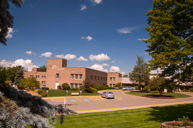 Front exterior of a brick senior living facility with a circular driveway, parked cars, and landscaped lawn under a blue sky.