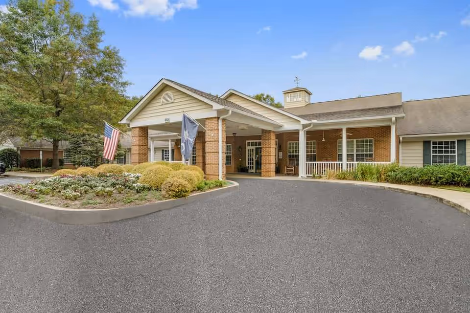 Front exterior view of a brick assisted living facility with a covered entrance, two flags, landscaped flower beds, and a clear blue sky.