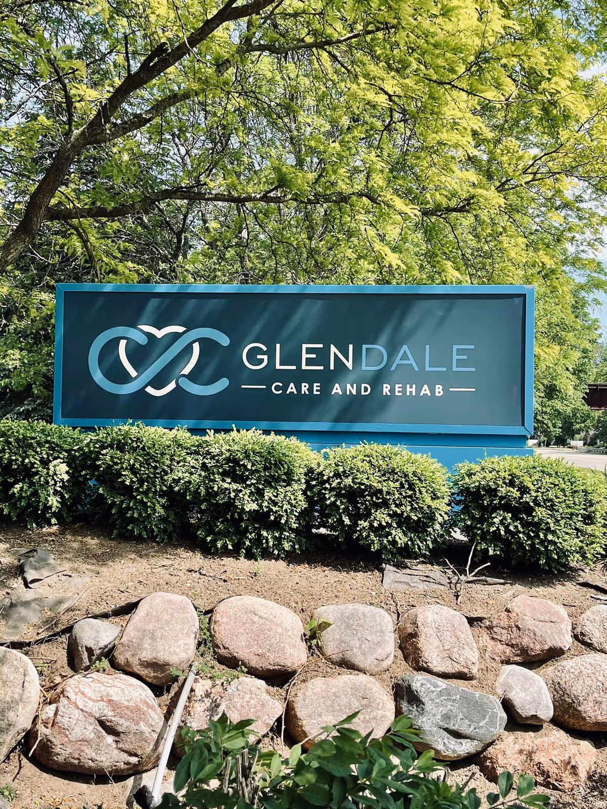 Outdoor teal sign reading 'GLENDALE CARE AND REHAB' set among shrubs, rocks, and trees.