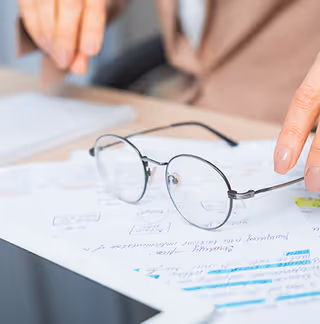 Glasses rest on documents with a notebook as hands point at papers on a desk.