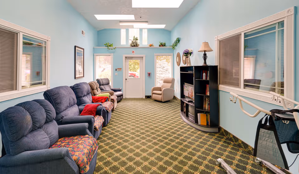 A bright, cozy common area with light blue walls and patterned carpet. The room features several cushioned recliner chairs lined up along the left wall, a beige armchair near the back door, and a black entertainment center with a TV and decorative items on the right. There are windows with blinds on both sides, a handrail on the right wall, and plants placed on a ledge above the door.