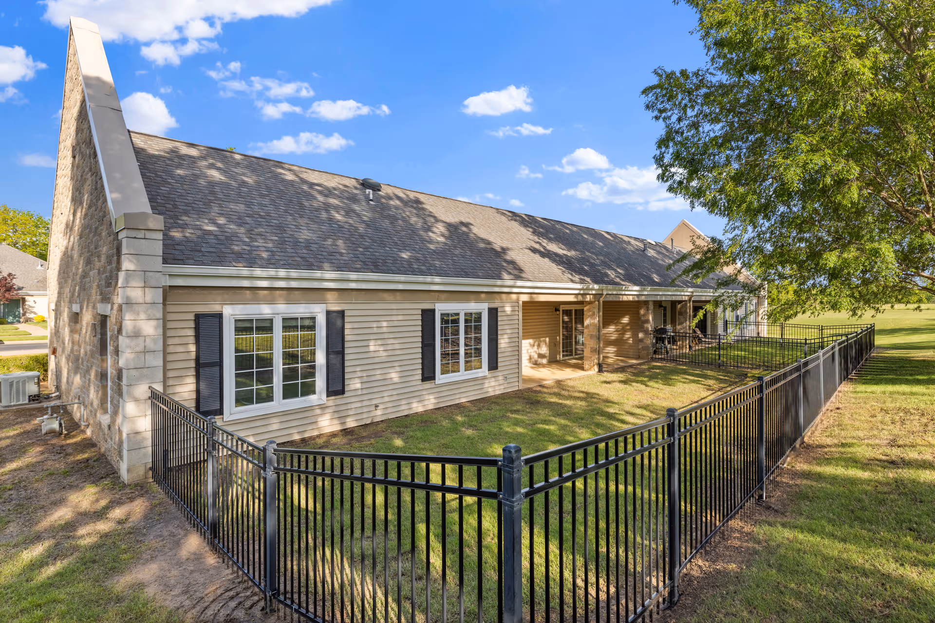Exterior view of a single-story building with beige siding and stone accents, featuring multiple windows with black shutters. The building has a sloped roof and a fenced grassy area in front, with a large tree on the right side under a blue sky with scattered clouds.