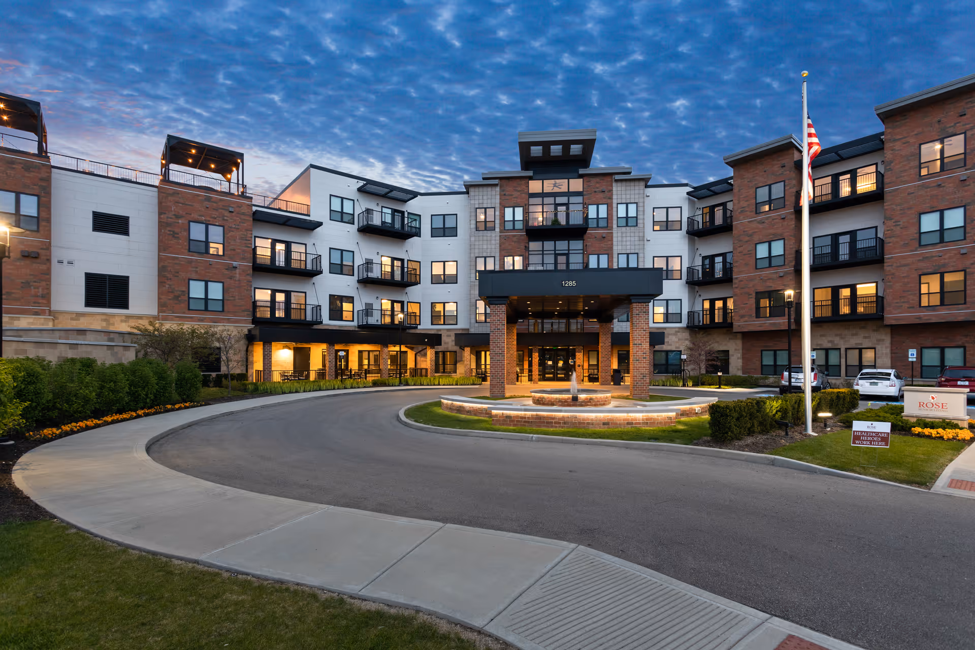 Front exterior of a multi-story senior living building at dusk with a circular driveway, fountain and American flag.