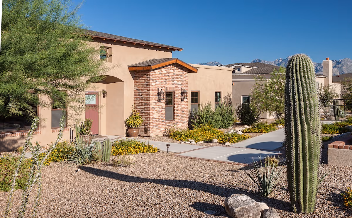 Front of a single-story desert-style building with brick and stucco facade, a walkway, gravel landscaping and a large cactus in the foreground.
