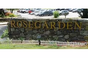 Stone wall with ROSEGARDEN sign and white fence, parking lot in background.