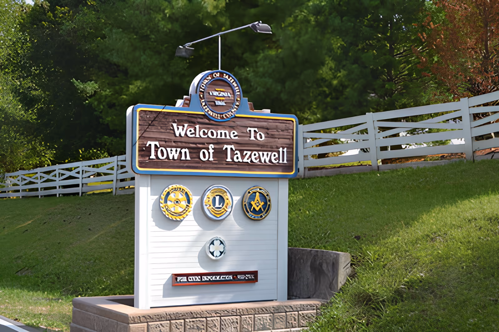 A welcome sign for the Town of Tazewell situated on a grassy area with a white fence and trees in the background. The sign features the text 'Welcome To Town of Tazewell' along with several circular emblems below the text.