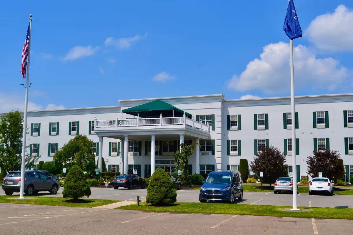 Front exterior of a white three-story senior living building with a columned entrance, parked cars, and flagpoles.