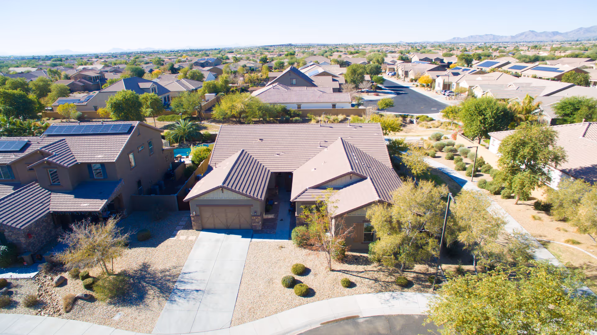 Aerial view of a suburban neighborhood centered on a single-story house with a driveway, surrounding homes, and desert landscaping.