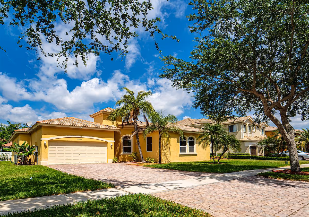A yellow, single-story senior living building with palm trees and a brick-paved driveway.
