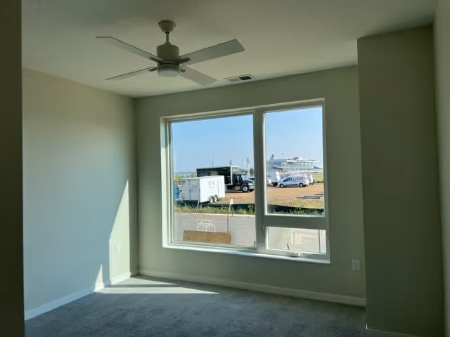 Empty room with light gray walls and carpeted floor, a ceiling fan with light fixture, and a large window showing an outdoor view of trucks and a fenced area.