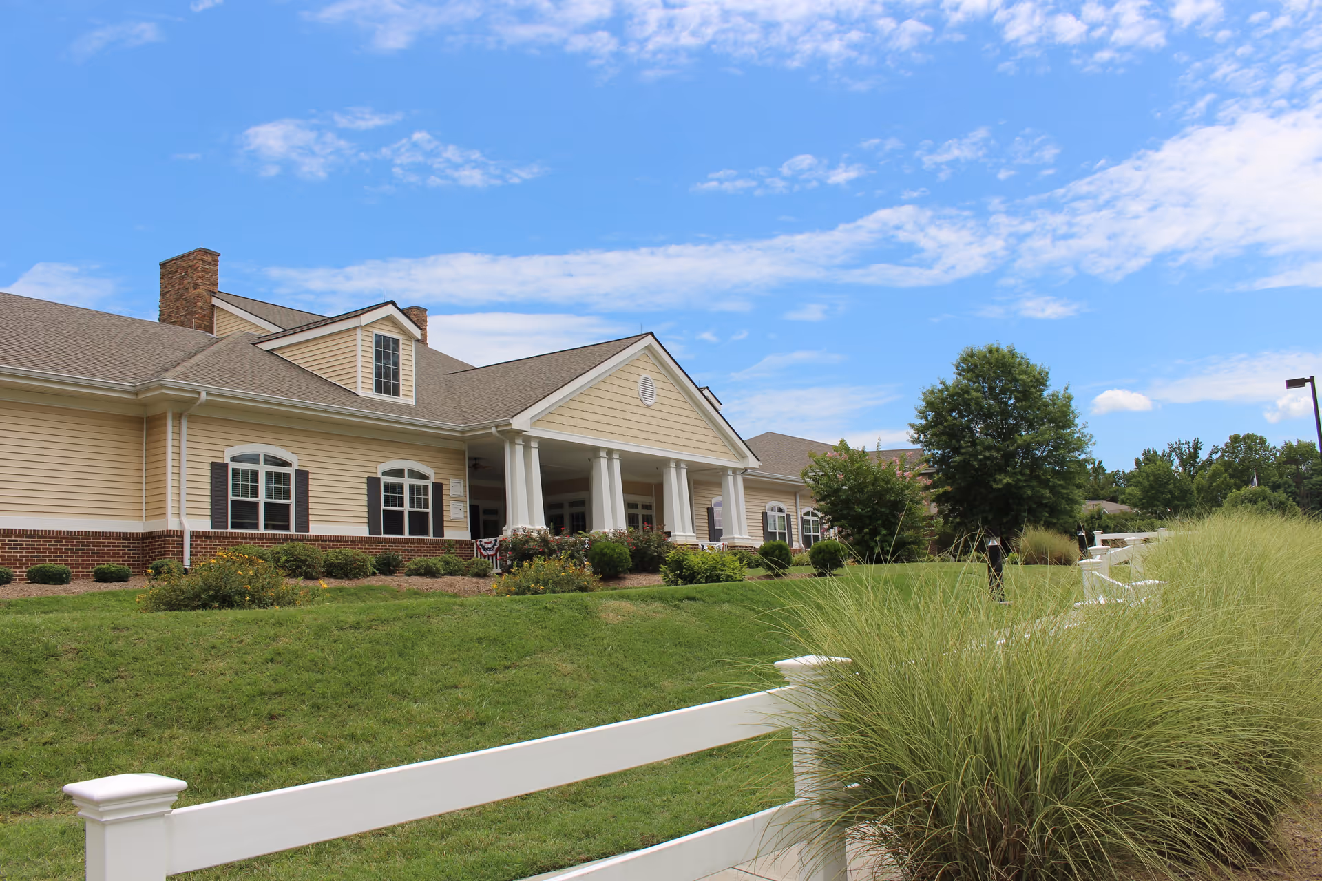 Exterior view of Piedmont Crossing Retirement Community building in Thomasville, NC, showing a beige and brick facade with white columns at the entrance, surrounded by green grass, bushes, and a white fence under a blue sky with scattered clouds.