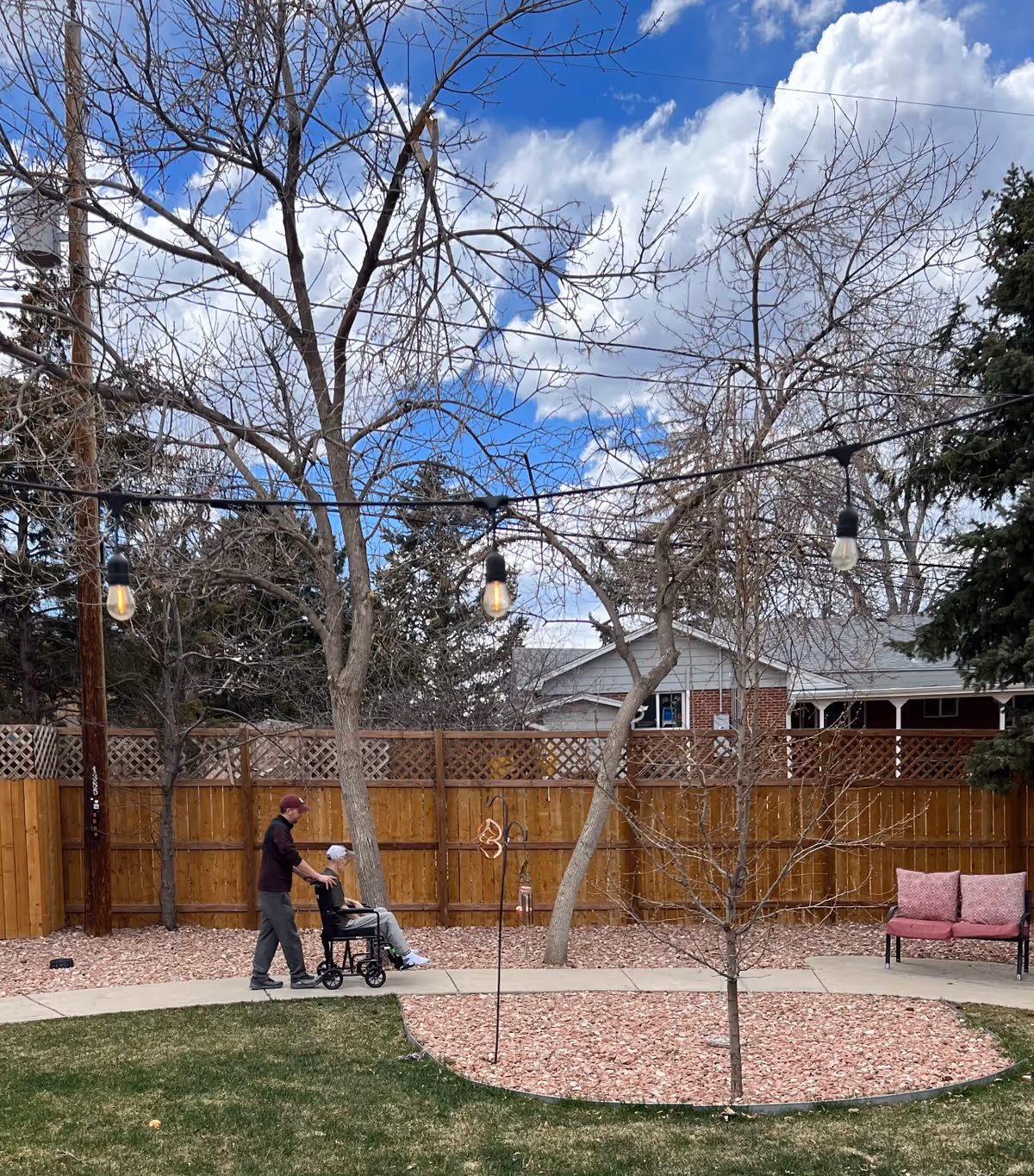 Caregiver pushes a resident in a wheelchair along an outdoor courtyard with string lights.