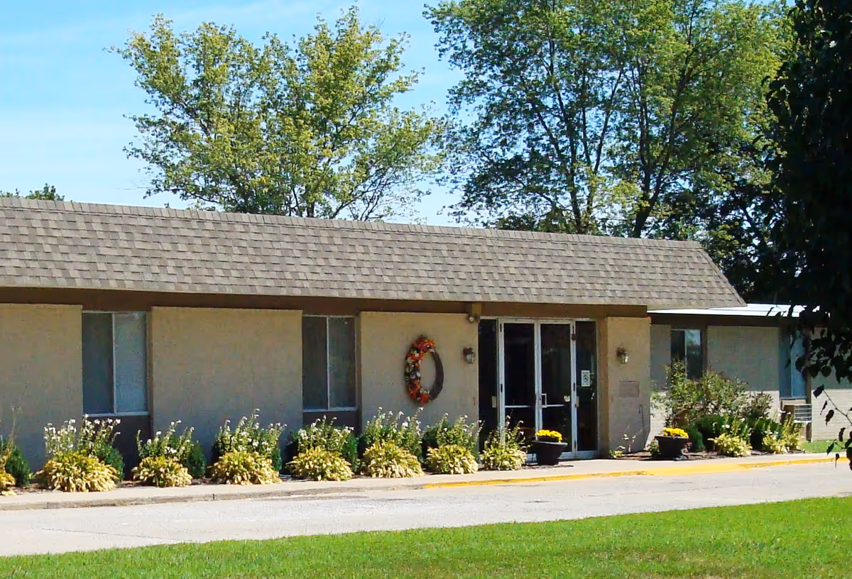 Single-story beige building façade showing the front entrance with double glass doors, a wreath, potted flowers, and landscaped beds.