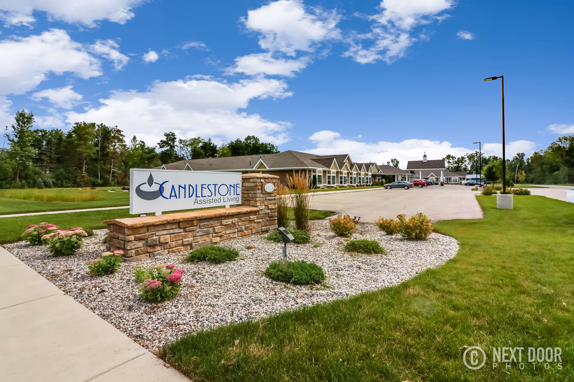 Exterior view of Candlestone Assisted Living facility with a stone sign in the foreground displaying the facility name. The building is single-story with a parking lot and several cars visible. The sky is partly cloudy and there is green grass and landscaping around the sign and building.