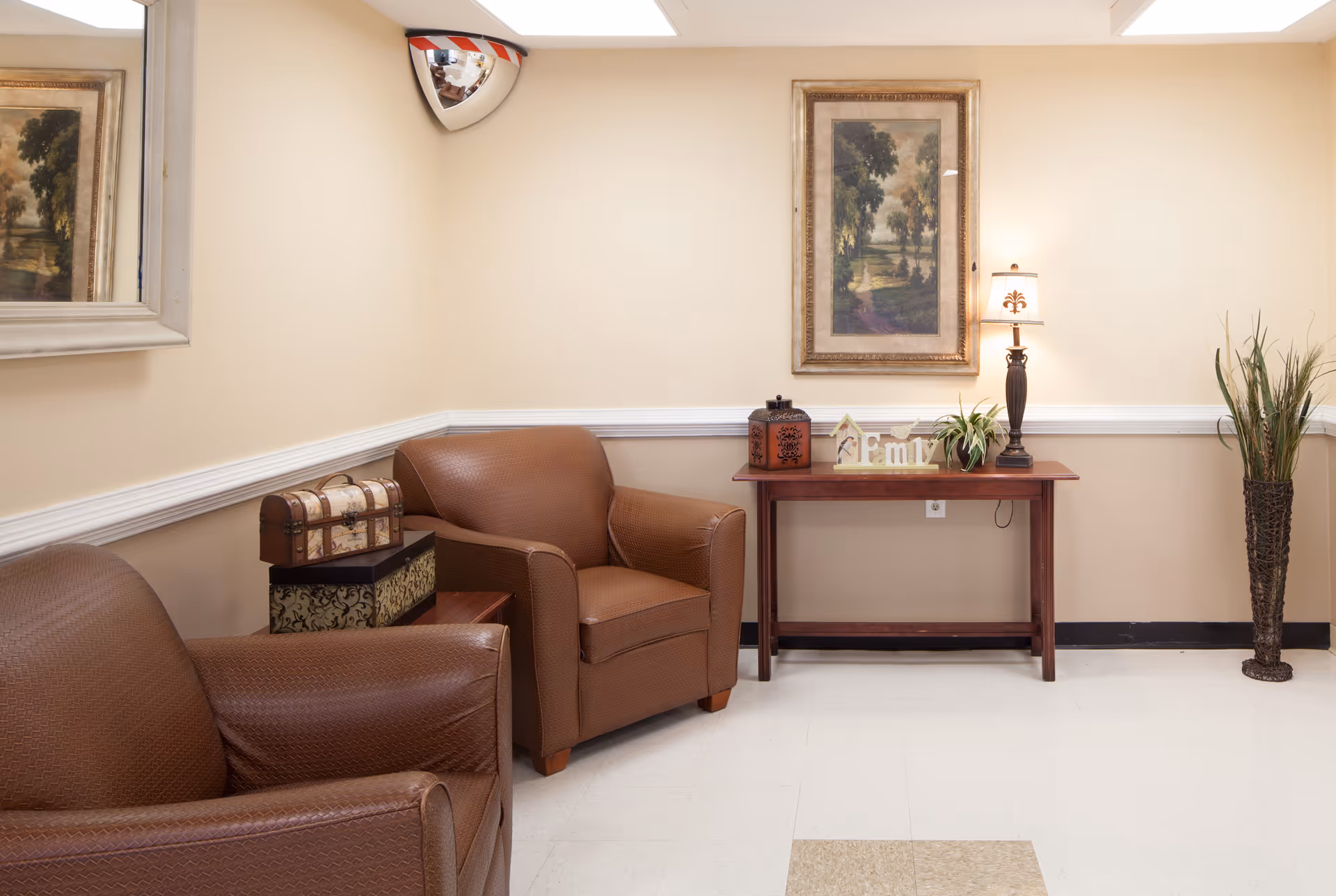 A cozy sitting area with two brown armchairs, a small wooden table with decorative boxes, a wooden console table with a lamp, plants, and a decorative 'Family' sign. A framed landscape painting hangs on the wall above the console table, and a convex security mirror is mounted in the corner near the ceiling.