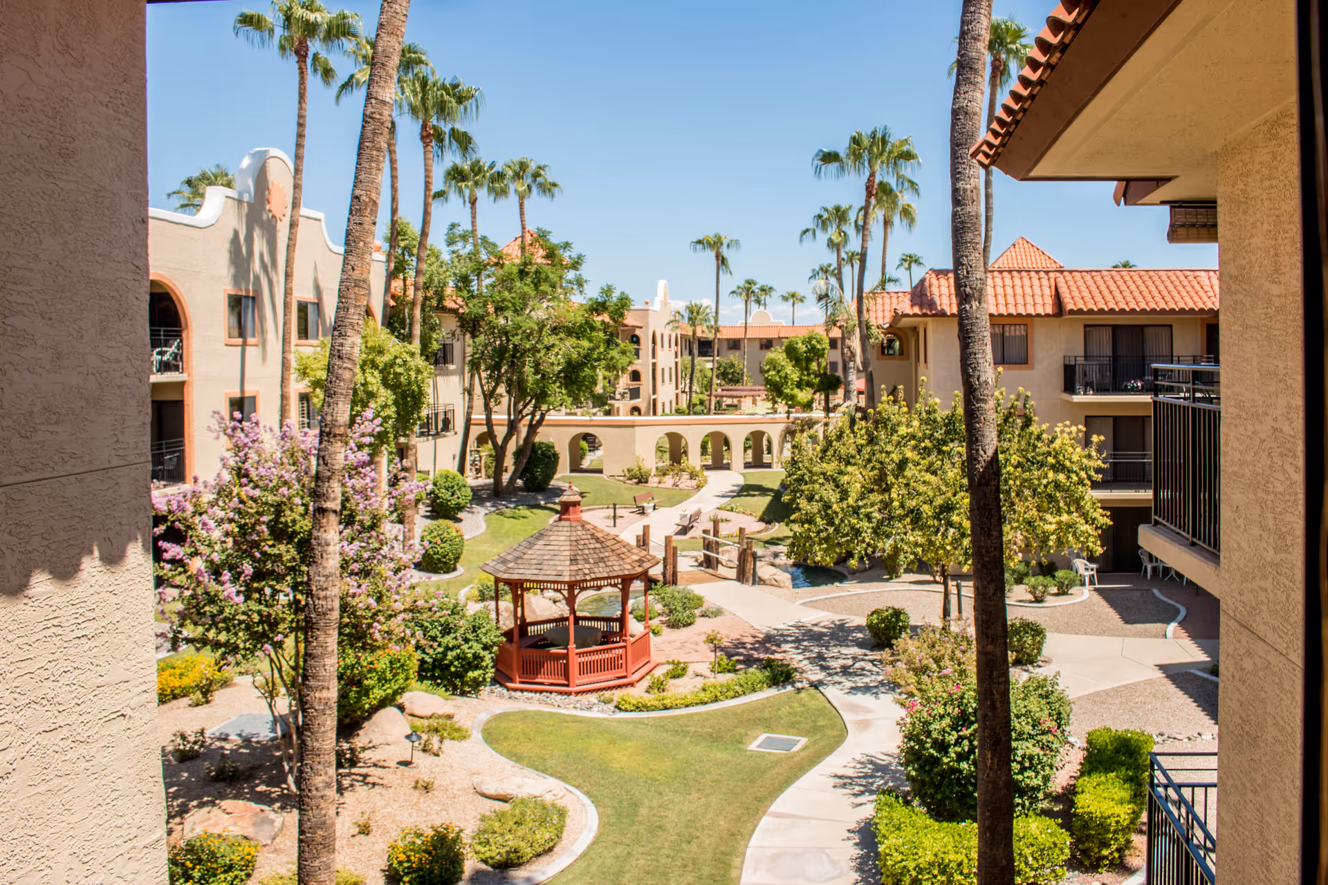 Sunny outdoor courtyard with palm trees, winding paths, a gazebo, and beige residential buildings.