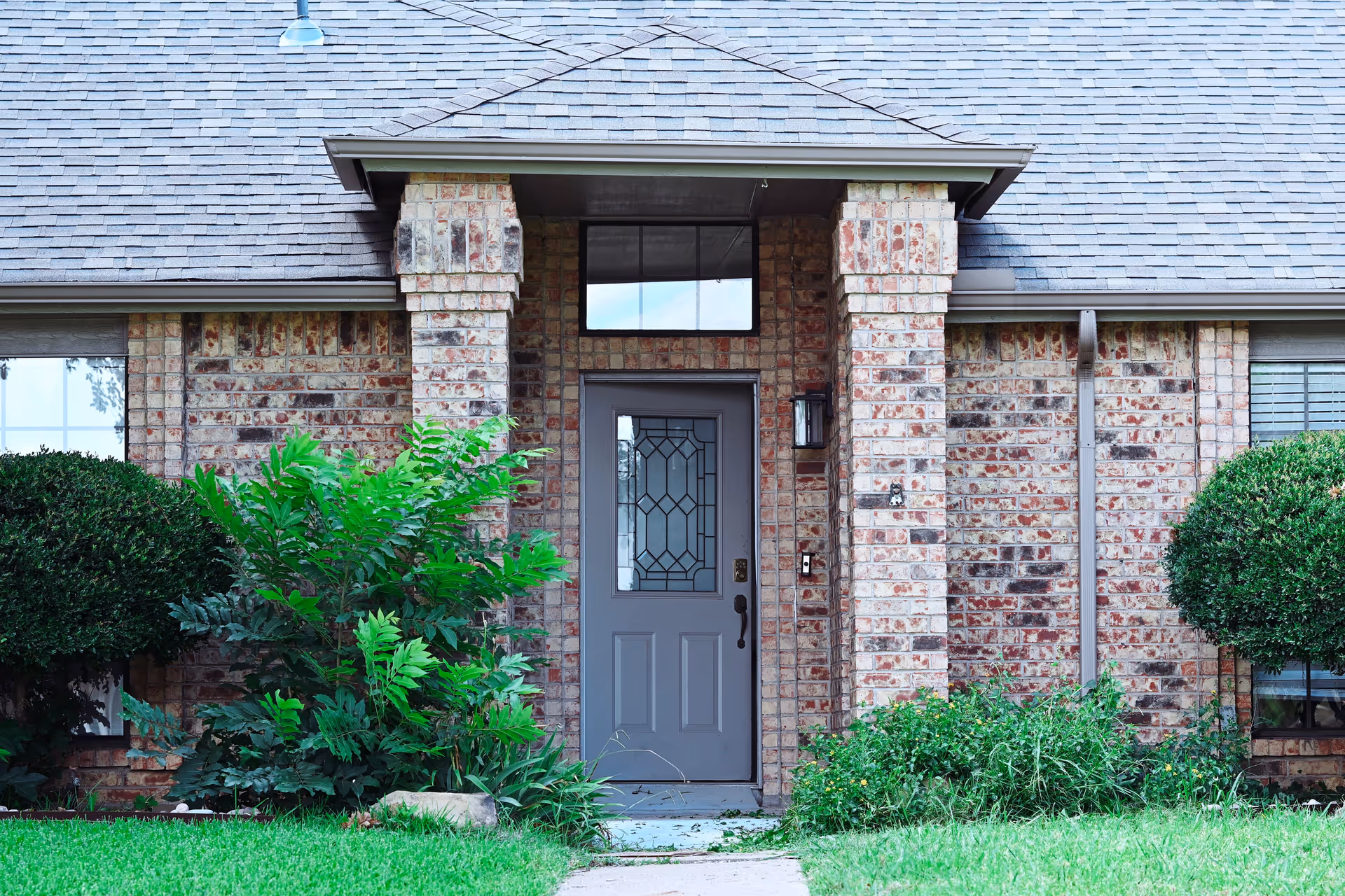 Front entrance of a brick residential building with shrubs and a gray door