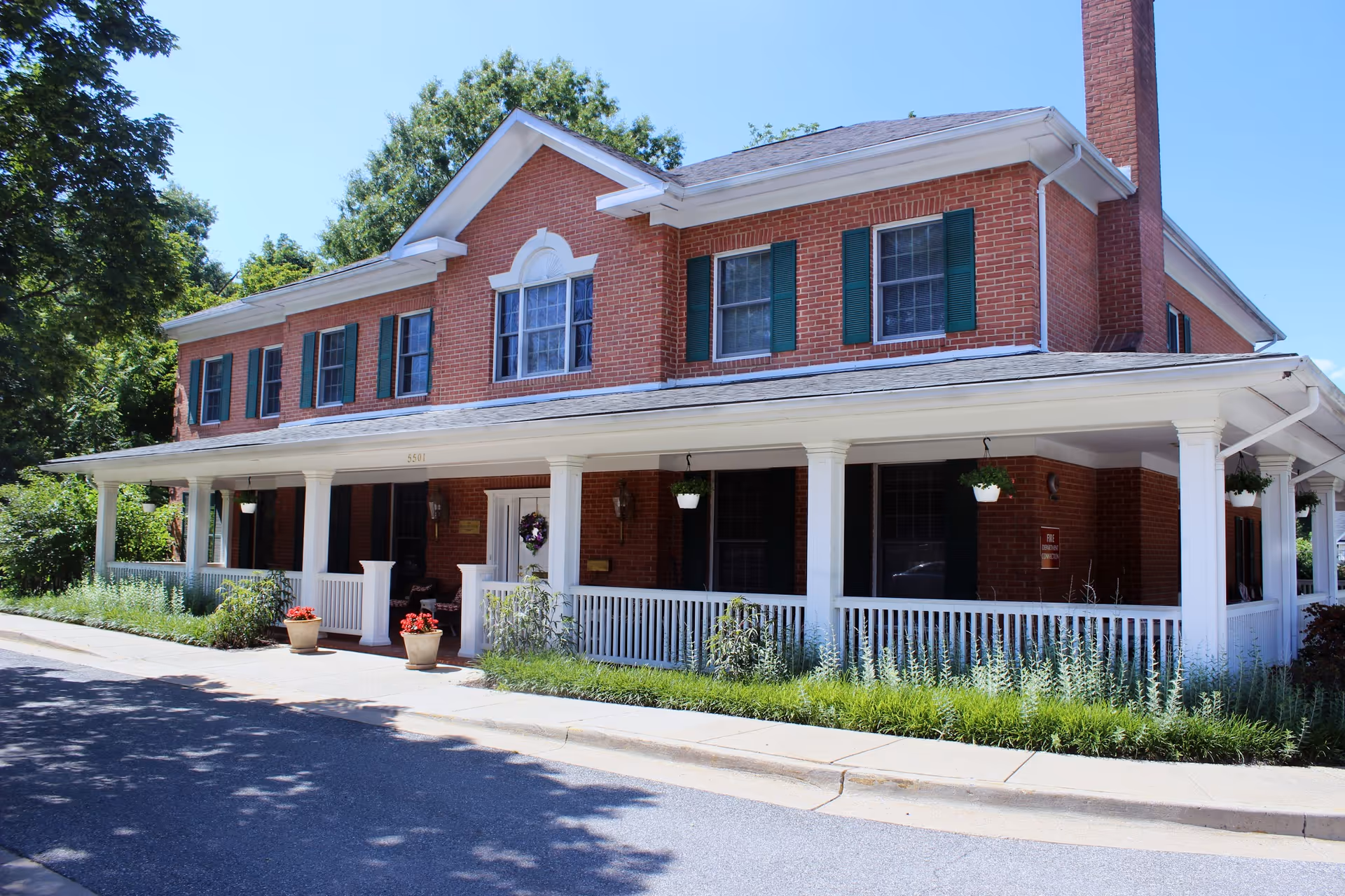 Two-story red brick house with a white wraparound porch, green shutters, hanging plants, and potted flowers in front.