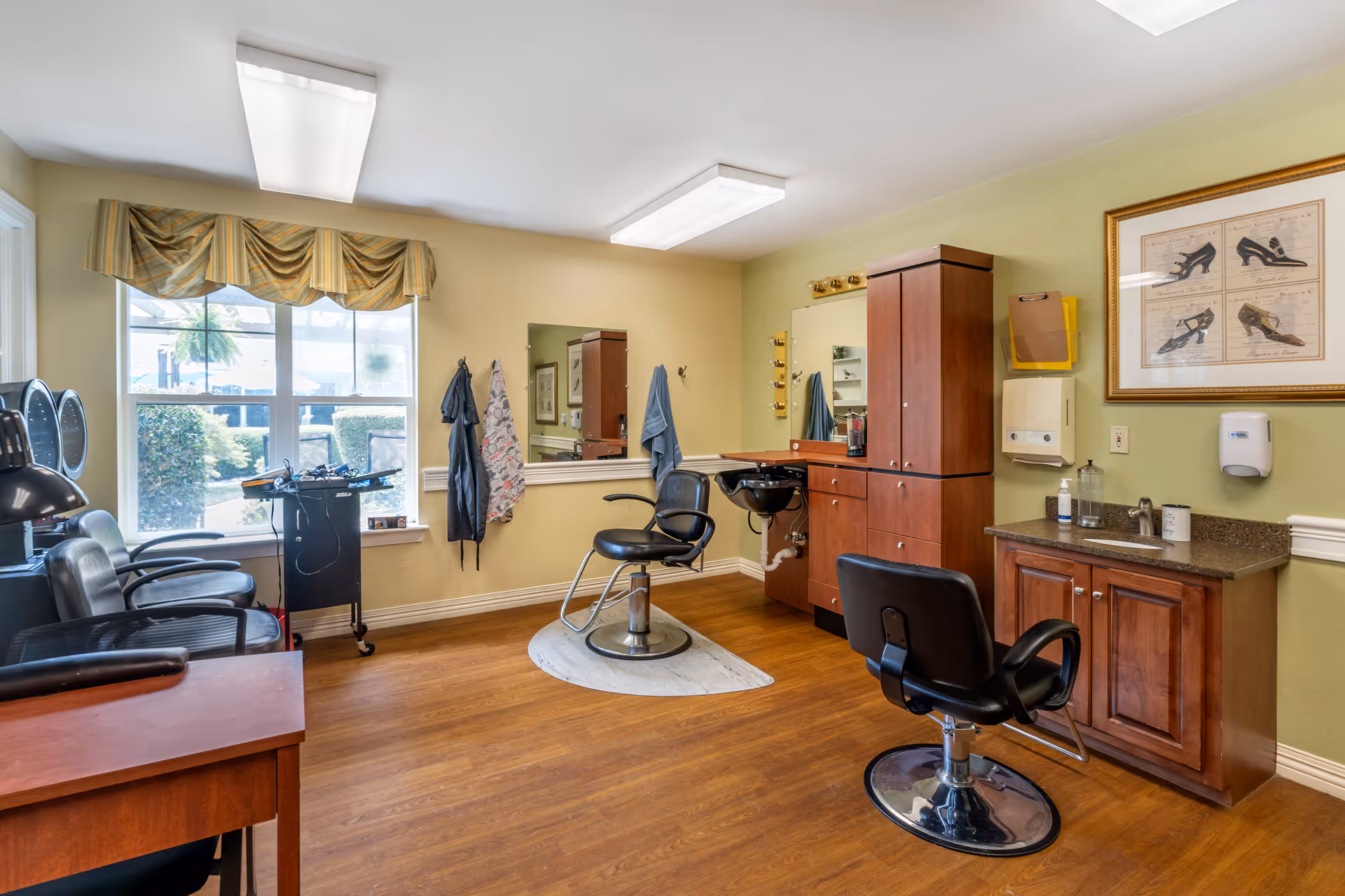 Interior view of a hair salon area in a senior living facility with wooden flooring, two black salon chairs, a hair washing station, a large mirror, and a window with a valance letting in natural light. There are cabinets, a sink, and hair care products on the counter, along with framed artwork on the wall.