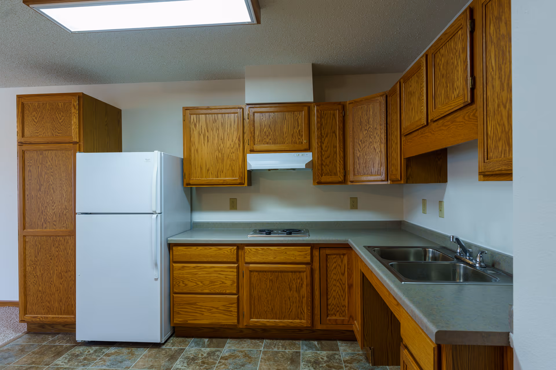 A kitchen with wooden cabinets, a white refrigerator, a double sink, a stovetop with a range hood, and a tiled floor. The countertops are gray, and the walls are white.