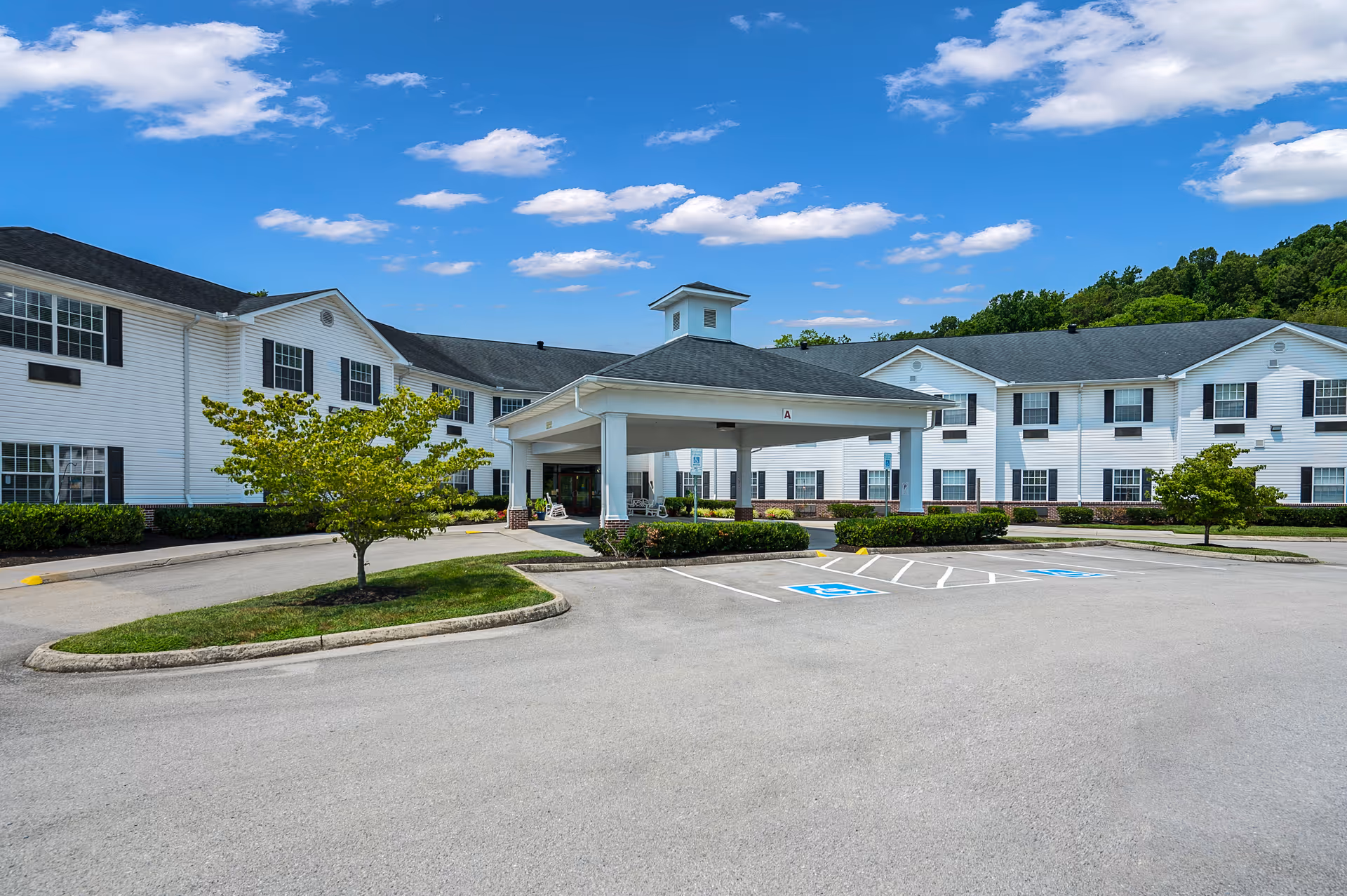 Exterior view of a senior living facility with a covered entrance and parking spaces, including handicapped spots. The building is white with black shutters and surrounded by greenery under a blue sky with scattered clouds.