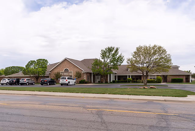 Single-story brick senior living facility with cars parked in front and trees nearby.