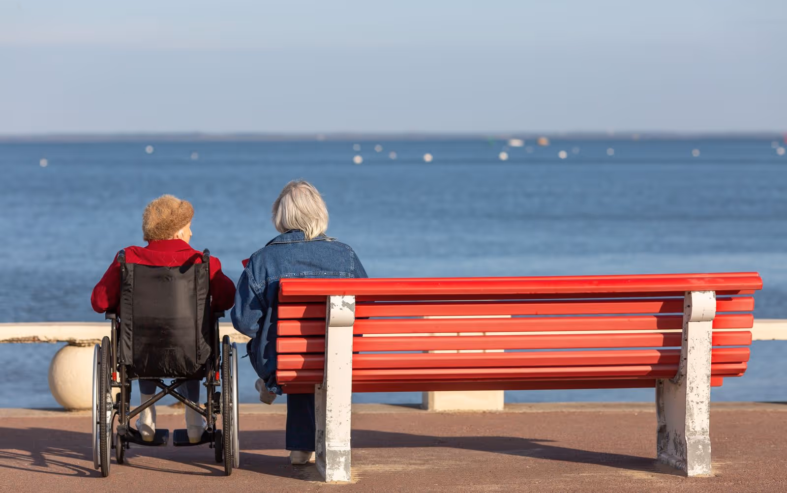 Two elderly people, one in a wheelchair, sit beside a red bench by the water.