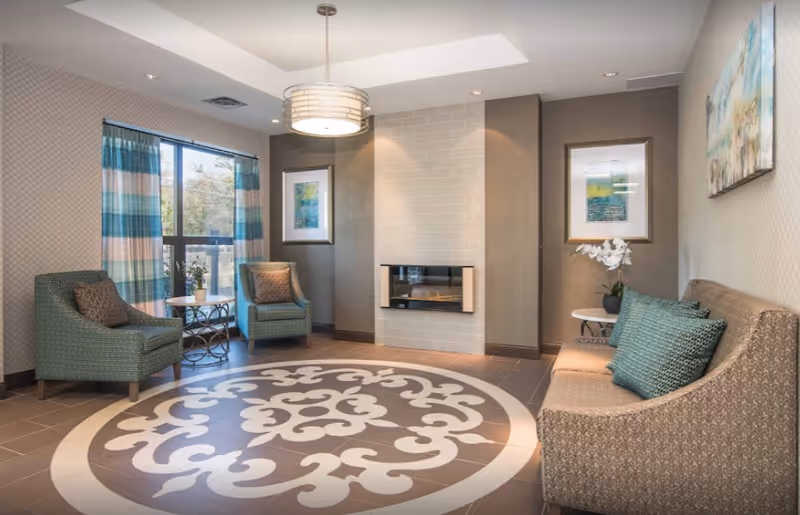 Communal living room with chairs, a sofa, a patterned round rug, and a wall-mounted fireplace under a pendant light.