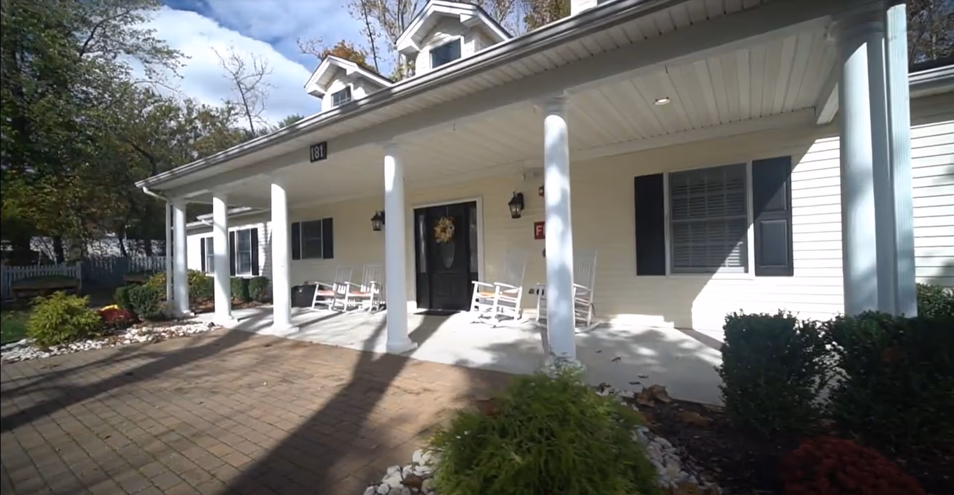 Front exterior view of a single-story building with white siding, black shutters, and a covered porch supported by white columns. The porch has several white rocking chairs and a decorative wreath on the black front door. The area is surrounded by landscaping with bushes and small trees under a partly cloudy sky.