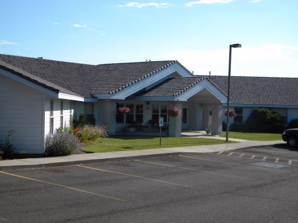 Exterior view of Chandler House facility showing a single-story building with a tiled roof, a covered entrance with hanging flower baskets, a small garden area, and an adjacent parking lot with marked spaces.