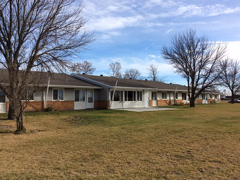 Single-story senior living building with a large grassy front yard and leafless trees under a partly cloudy sky.