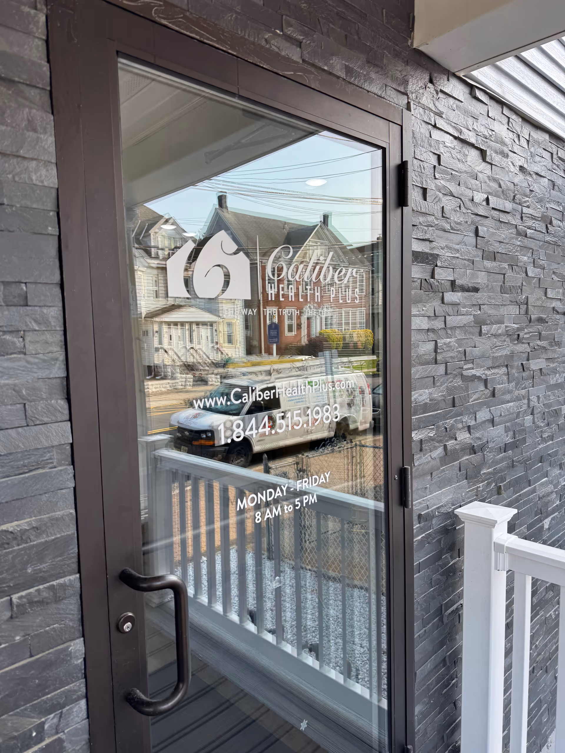 Glass entrance door with Caliber Health Plus logo on a stone exterior wall, reflecting street.