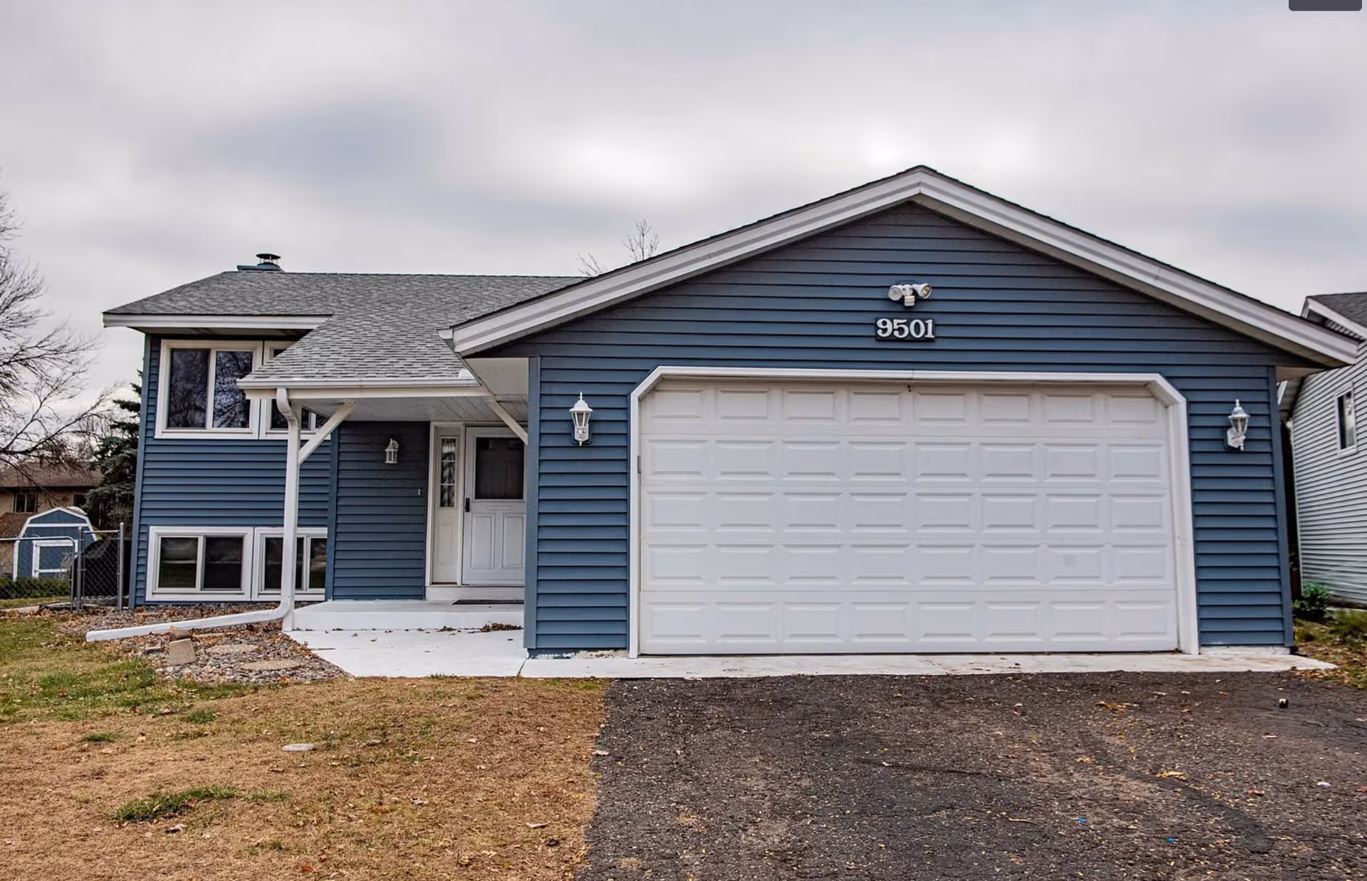 Blue single-family home with a large white garage door and driveway under cloudy sky.