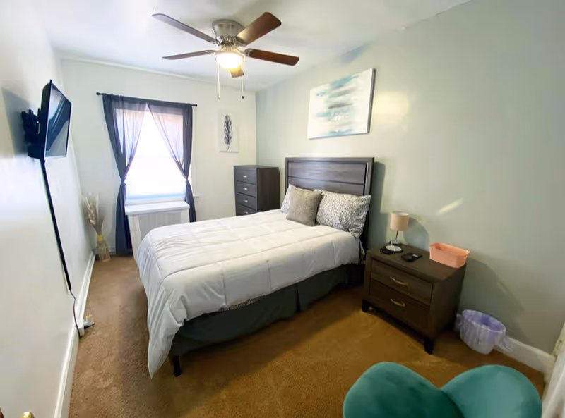 Bedroom in a senior living facility with bed, dresser, window, and ceiling fan.