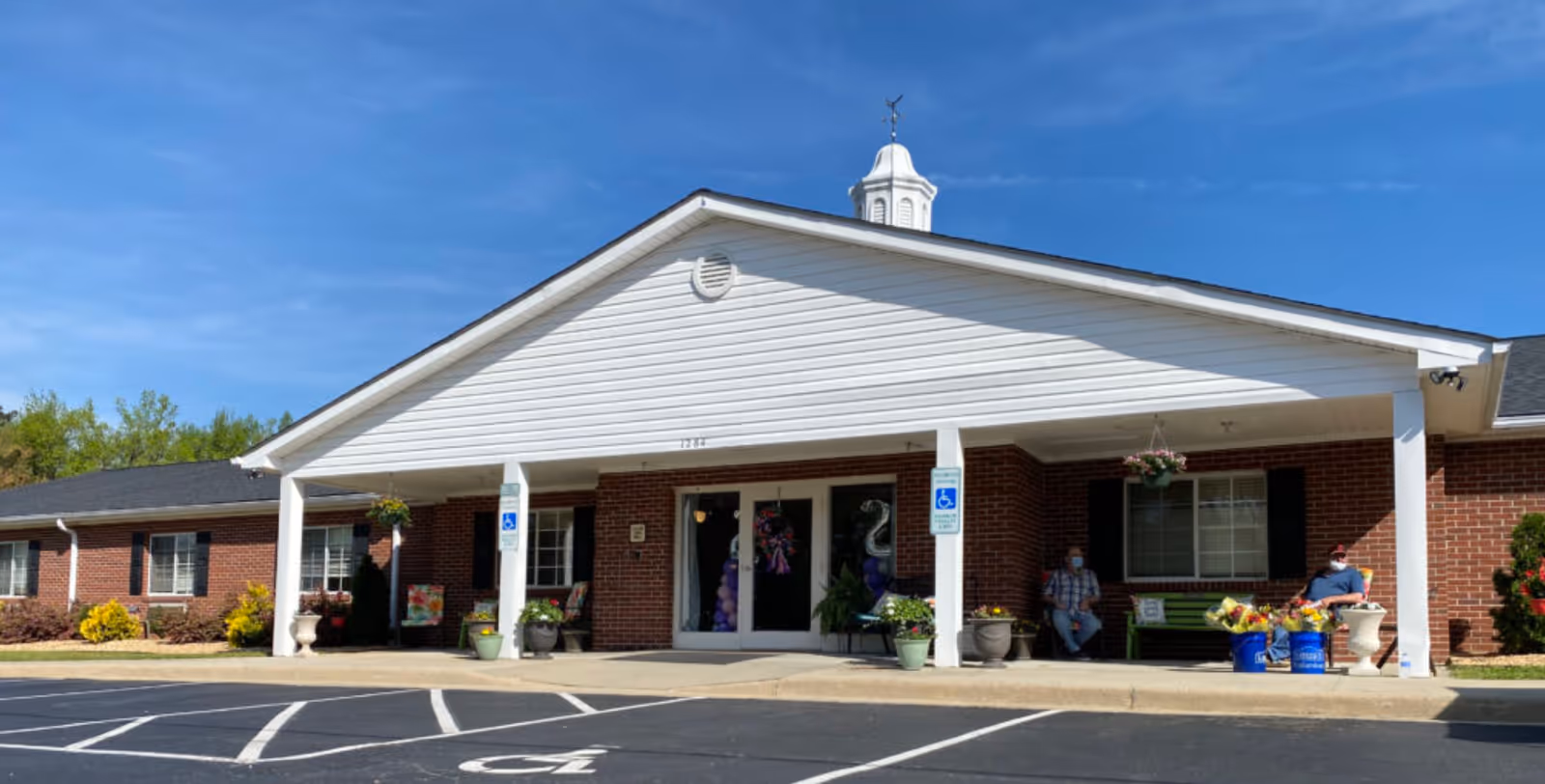 Front entrance of a single-story brick senior living building with a white covered portico, potted plants, handicap parking, and two people seated on the porch.