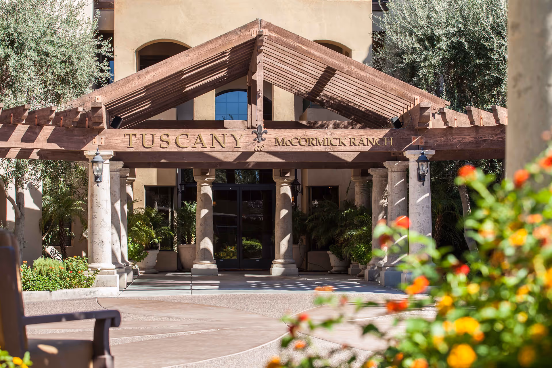 Entrance to Tuscany at McCormick Ranch featuring a wooden pergola with the facility name engraved, stone columns, and surrounding greenery with flowers in the foreground.