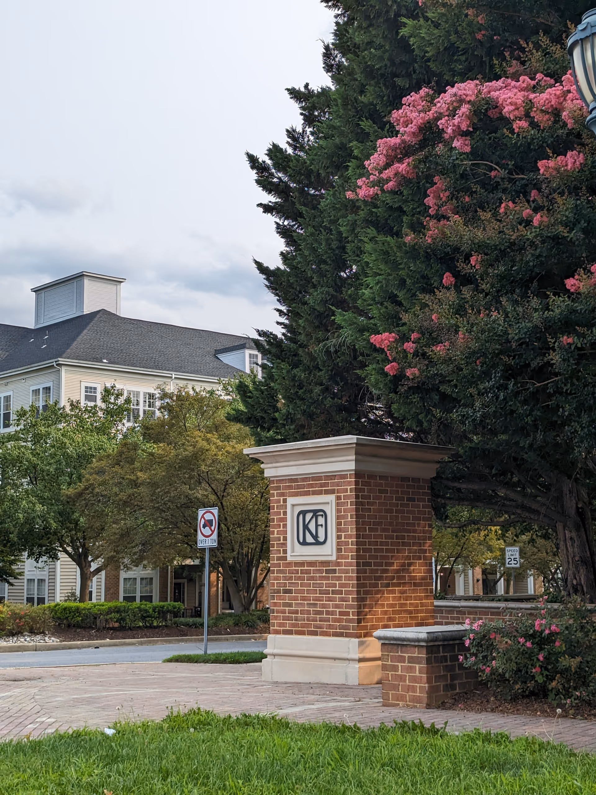 Brick entrance pillar with a sign, trees, and a building in the background.